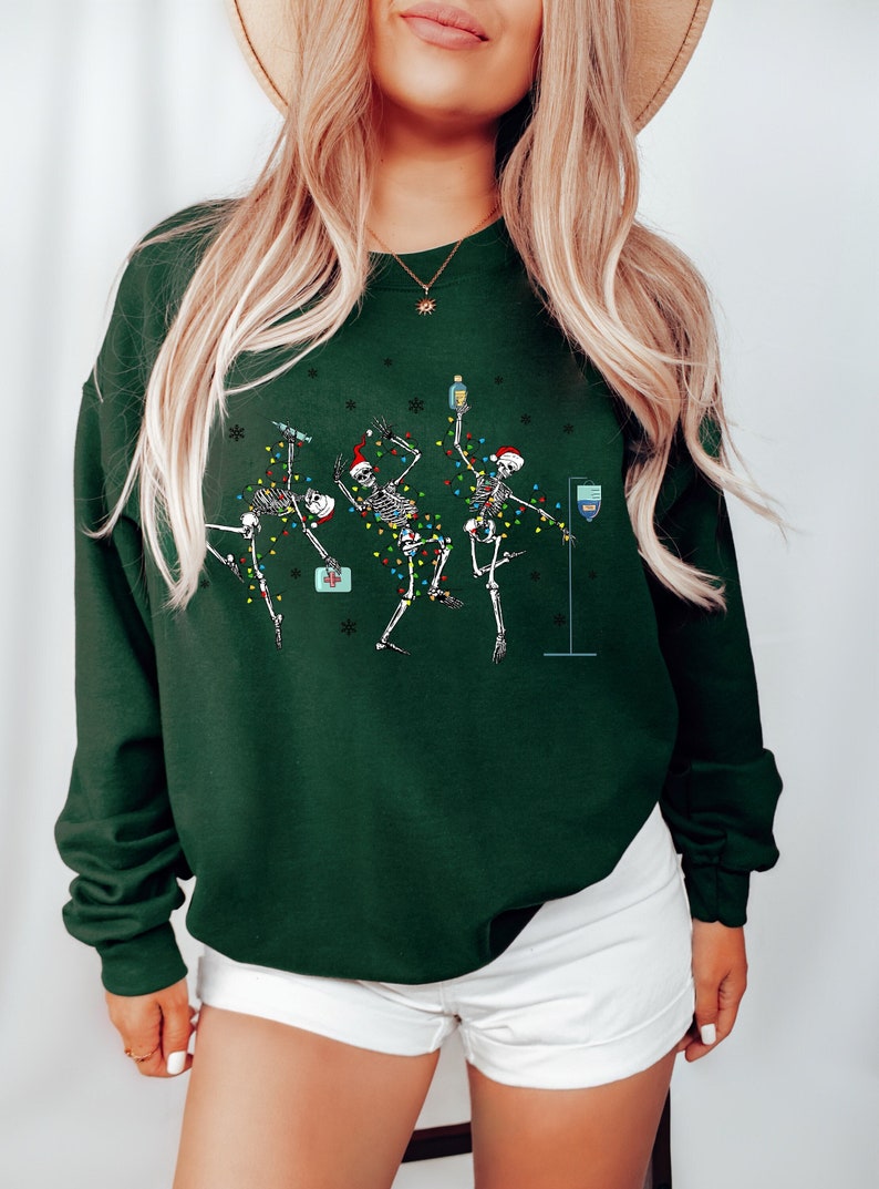 May include: A green sweatshirt with a festive design featuring three skeletons dressed in Santa hats and Christmas lights. The skeletons are holding drinks and a first aid kit.