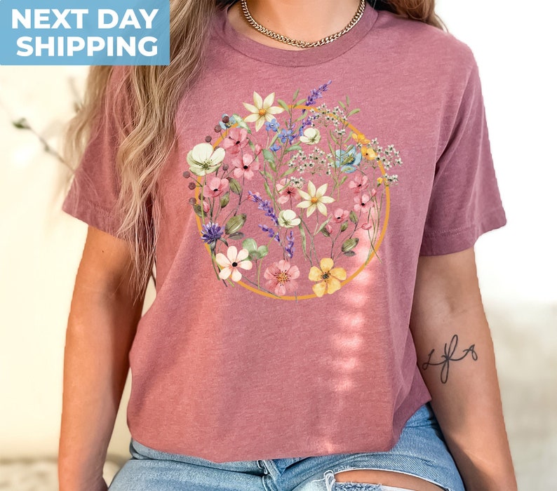 May include: A mauve colored t-shirt with a circle design featuring a watercolor floral pattern in shades of pink, yellow, blue, and green.