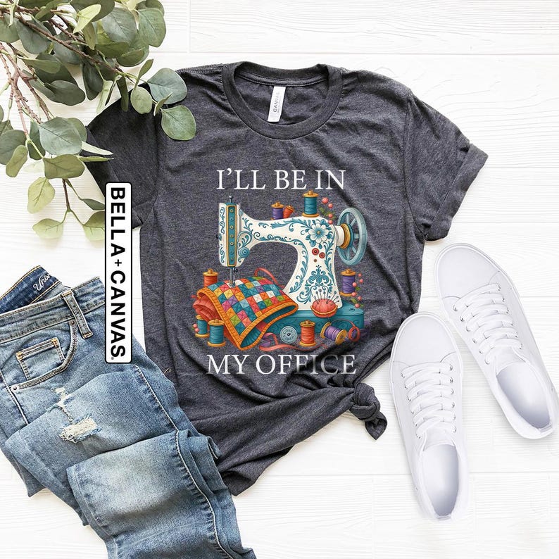 May include: Dark gray t-shirt with a sewing machine graphic and the text "I'll Be In My Office." The graphic includes a sewing machine, colorful spools of thread, and a quilt. White sneakers and blue jeans are also in the image.