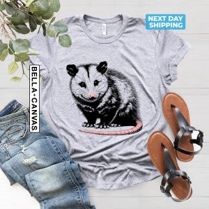 K&ouml;nnte beinhalten: Graues T-Shirt mit einer schwarz-wei&szlig;en Illustration eines Opossums. Das Opossum sitzt auf seinen Hinterbeinen mit dem Schwanz um den K&ouml;rper gewickelt. Der Text "BELLA + CANVAS" ist auf der linken Seite des Shirts gedruckt.