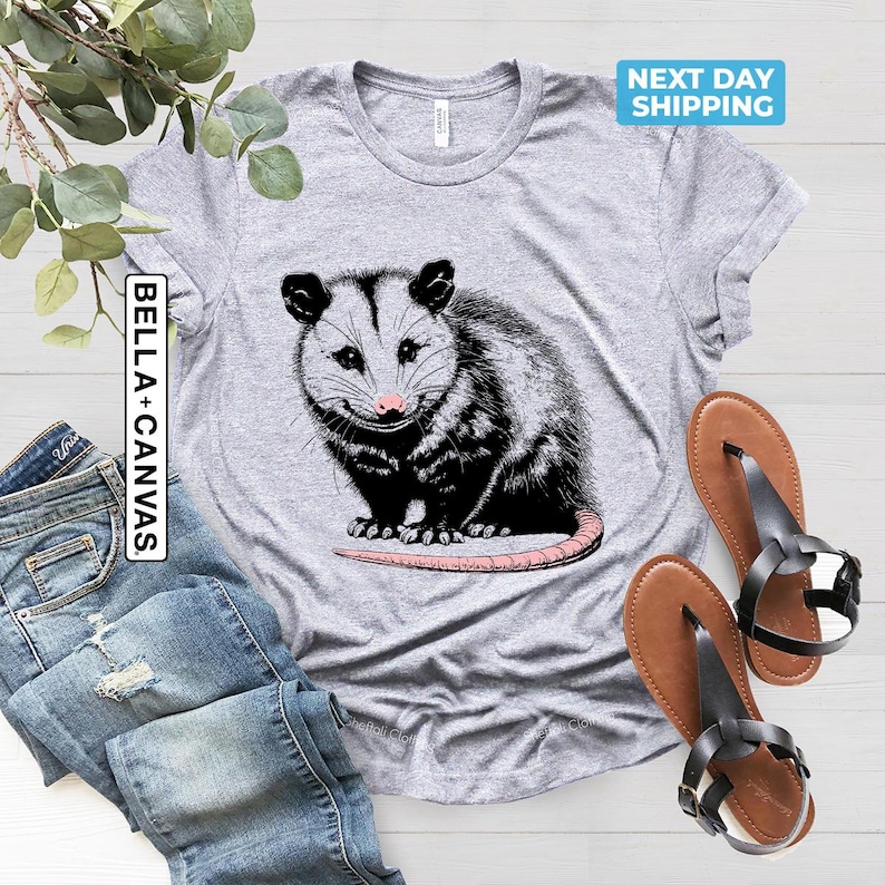 K&ouml;nnte beinhalten: Graues T-Shirt mit einer schwarz-wei&szlig;en Illustration eines Opossums. Das Opossum sitzt auf seinen Hinterbeinen mit dem Schwanz um den K&ouml;rper gewickelt. Der Text "BELLA + CANVAS" ist auf der linken Seite des Shirts gedruckt.
