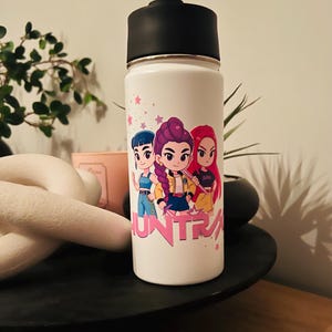 May include: White insulated water bottle with a black lid and gold rim. The bottle features a cartoon graphic of three girls with colourful hair and the word "HUNTRA" in pink. Pink stars are scattered around the design.