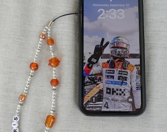 以下が含まれることがあります： 銀色のチェーンとオレンジ色のビーズでできた携帯電話のチャームで、白い文字で「NORRIS」と綴られています。チャームは、レーシングドライバーの写真が映し出された黒いスマートフォンに取り付けられています。