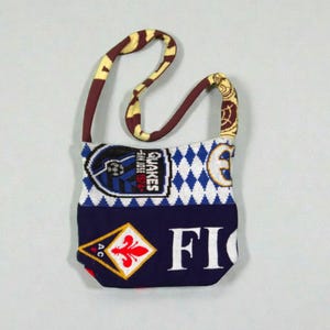 May include: A small, fabric shoulder bag with a patterned strap. The bag features a patchwork design with a soccer team logo, diamond pattern, and the word "FIO" in white letters. The strap is brown and yellow.