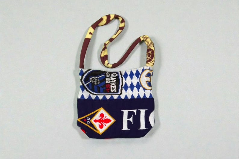 May include: A small, fabric shoulder bag with a patterned strap. The bag features a patchwork design with a soccer team logo, diamond pattern, and the word "FIO" in white letters. The strap is brown and yellow.