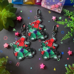 Puede incluir: Tres llaveros de acrílico que muestran un personaje de dibujos animados con cabeza roja, cuerpo verde y una cola en forma de cable de alimentación negro. Cada llavero tiene un cierre de mosquetón de metal plateado en la parte superior.