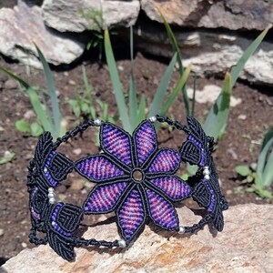 May include: A handmade macrame bracelet featuring a large, central flower design. The flower has purple and pink petals with black outlines. The bracelet is black with silver beads and is displayed on a stone surface.