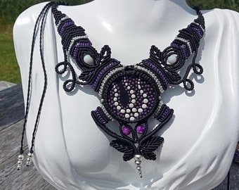 May include: A handmade macrame necklace featuring a central eye design in black, purple, and white. The necklace has a detailed, symmetrical pattern with decorative elements and a black cord. The necklace is displayed on a white mannequin bust.