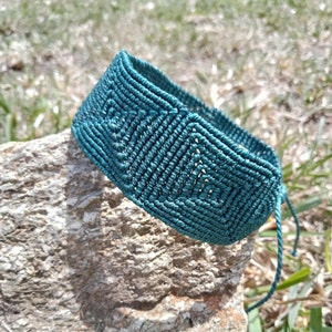 May include: Teal macrame bracelet with a geometric pattern. The bracelet is made of woven cord and is resting on a textured rock. The bracelet has a square design and is tied with teal cords.