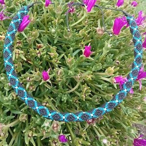 Puede incluir: Un collar de macramé hecho a mano con un patrón geométrico repetitivo en turquesa, gris y blanco. El collar se muestra sobre un fondo de follaje verde y flores moradas, mostrando su intrincado diseño.
