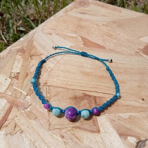 May include: A turquoise macrame bracelet with a sliding knot closure. The bracelet features a series of beads in shades of purple and turquoise. The bracelet is displayed on a wooden surface.