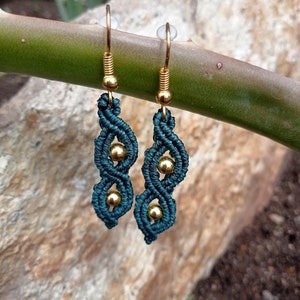 May include: Teal macrame earrings with gold-colored beads and ear wires. The earrings have a woven design with three gold beads. The earrings are hanging against a blurred background of a green stem and a rock.