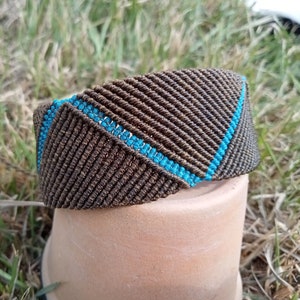 Può includere: Un braccialetto macramè fatto a mano nei toni del marrone e turchese. L'ampio braccialetto presenta un motivo diagonale marrone con una striscia turchese. Il braccialetto è esposto su un vaso di terracotta su erba verde.
