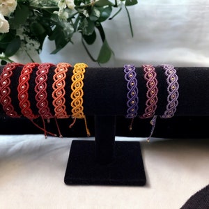 May include: A collection of handmade macrame bracelets in various colors, including red, orange, yellow, and purple. Each bracelet features a woven design with silver or gold-colored beads. The bracelets are displayed on a black velvet stand.