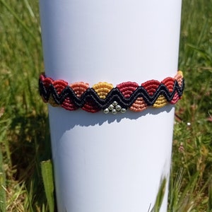 以下が含まれることがあります： 赤、オレンジ、黄色、黒のジグザグ模様のマクラメブレスレット。ブレスレットの中央には小さな銀色のビーズクラスターがあります。白い円筒形の物体の上に、緑の草を背景に表示されています。