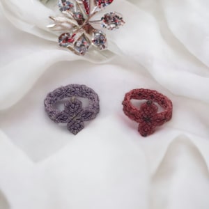Peut inclure: Deux anneaux en crochet, un violet et un bordeaux, avec un motif floral. Au-dessus, une broche en forme de feuille argentée avec des accents rouges, blancs et bleus. Les objets sont posés sur un tissu de soie blanc, avec une branche de conifère en arrière-plan.