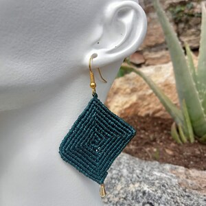 Può includere: Orecchini in macramè color turchese con un design a forma di diamante. Gli orecchini pendono da ganci color oro e presentano una piccola perla dorata sul fondo. Sono esposti su una testa di manichino bianca.