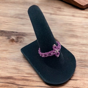 Peut inclure: Une bague en macramé faite à la main, dans des tons de violet et de blanc, présentée sur un présentoir noir. La bague présente un motif tissé, mettant en valeur la technique de nouage complexe. Le fond est une surface en bois.