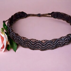 May include: A dark brown macrame headband with a zig-zag pattern. A pink rose with green leaves is placed next to the headband on a pink background. The headband is adjustable.