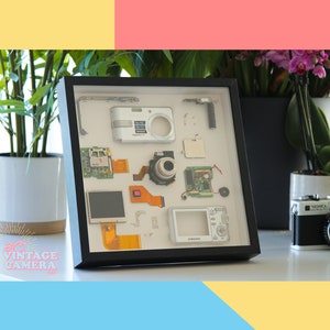 Puede incluir: Una caja de sombra enmarcada en negro con un fondo blanco muestra piezas desmontadas de una cámara digital. Las piezas incluyen una lente, placas de circuito, una pantalla y un cuerpo de cámara. La caja de sombra está sentada sobre una superficie blanca.