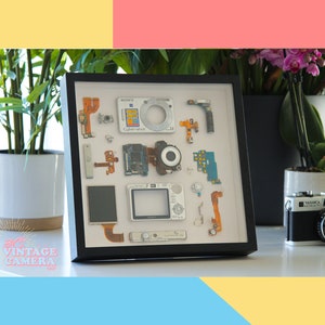 Puede incluir: Una vitrina enmarcada de piezas desmontadas de una cámara digital Sony Cyber-shot. Las piezas están dispuestas sobre un fondo blanco e incluyen la lente, la placa de circuito, los botones y otros componentes.