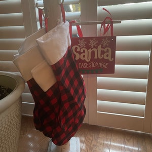 May include: A white wooden stand with a red and black plaid stocking hanging from it. The stand has a sign that says "Santa Please Stop Here" with snowflakes.