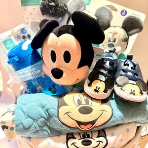 Op de afbeelding: Een babycadeaupakket gevuld met Disney-artikelen. Inclusief een Mickey Mouse-knuffel, blauwe babyschoenen, een pop-up rietjesbeker en babykleding met Mickey Mouse-ontwerpen. De mand is wit en geweven.