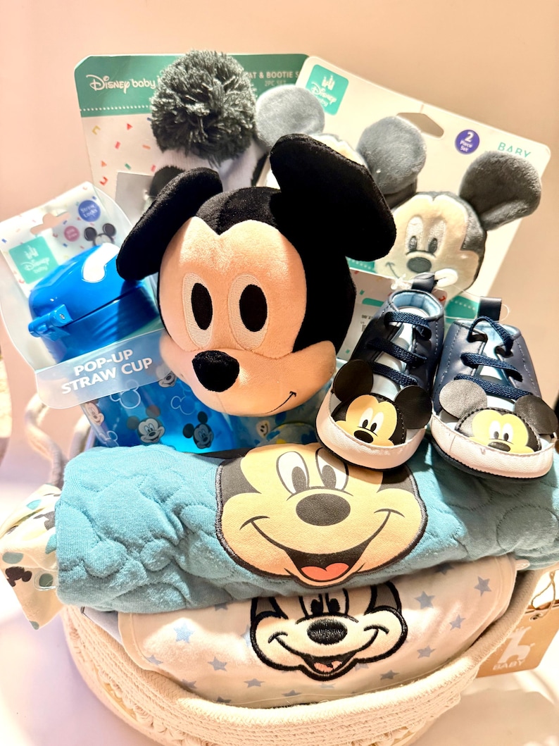 Op de afbeelding: Een babycadeaupakket gevuld met Disney-artikelen. Inclusief een Mickey Mouse-knuffel, blauwe babyschoenen, een pop-up rietjesbeker en babykleding met Mickey Mouse-ontwerpen. De mand is wit en geweven.
