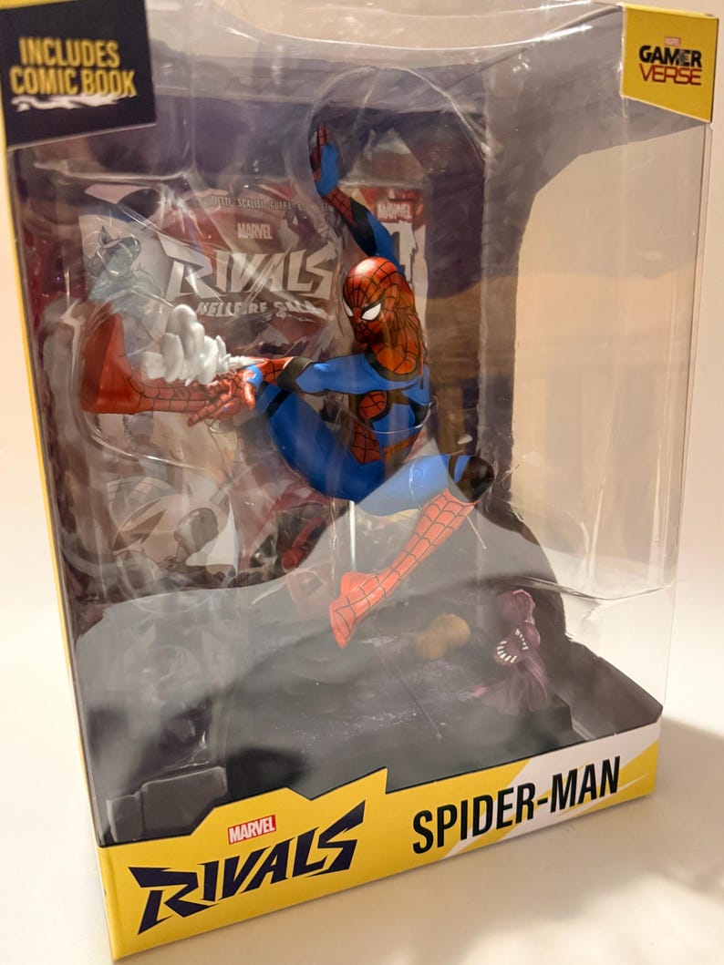 May include: A Spider-Man action figure in a clear plastic display case. The figure is in a dynamic pose, wearing a red and blue suit. The packaging includes the text "Marvel Rivals" and "Spider-Man". The box also says "Includes Comic Book" and "Gamer Verse".