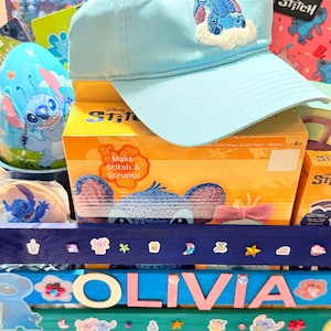 K&ouml;nnte beinhalten: Hellblaue Baseballkappe mit Stitch-Stickerei, ein Stitch-Rucksack und verschiedene Stitch-Geschenkartikel. Die Artikel sind auf einer farbenfrohen, dekorierten Oberfl&auml;che mit dem Namen "OLIVIA" angeordnet.