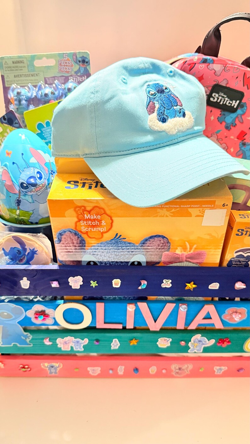 K&ouml;nnte beinhalten: Hellblaue Baseballkappe mit Stitch-Stickerei, ein Stitch-Rucksack und verschiedene Stitch-Geschenkartikel. Die Artikel sind auf einer farbenfrohen, dekorierten Oberfl&auml;che mit dem Namen "OLIVIA" angeordnet.