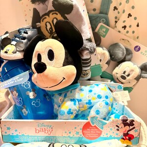 Op de afbeelding: Een Disney baby cadeaupakket met Mickey Mouse items. Het bevat een pluche speelgoed, een blauwe pop-up rietjesbeker, een hoed, schoenen, een slab en een onesie. De onesie is lichtblauw met gele en blauwe Mickey Mouse patronen.