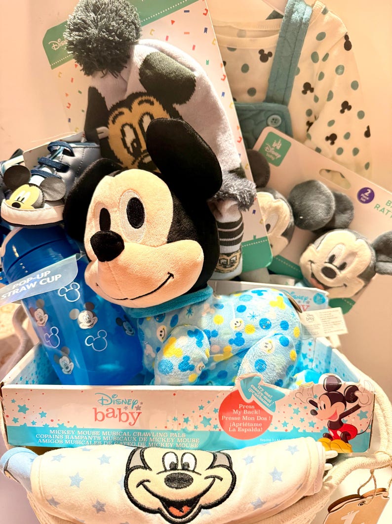 Op de afbeelding: Een Disney baby cadeaupakket met Mickey Mouse items. Het bevat een pluche speelgoed, een blauwe pop-up rietjesbeker, een hoed, schoenen, een slab en een onesie. De onesie is lichtblauw met gele en blauwe Mickey Mouse patronen.