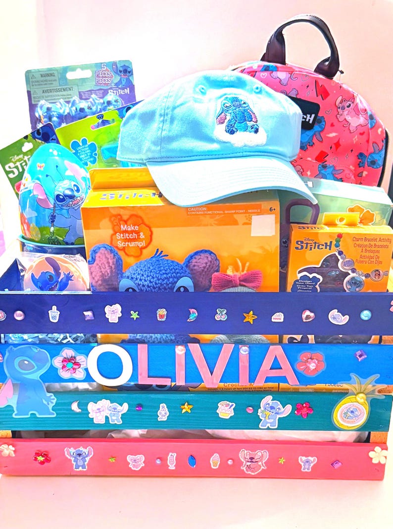 K&ouml;nnte beinhalten: Eine hellblaue Baseballkappe mit einem Stitch-Design liegt auf einer Holzkiste voller Stitch-Artikel. Die Kiste ist mit Aufklebern und dem Namen "OLIVIA" verziert. Weitere Artikel sind ein rosa Rucksack und Bastelsets.