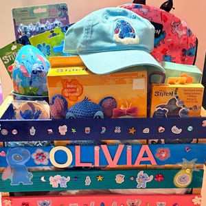 K&ouml;nnte beinhalten: Eine Holzkiste gef&uuml;llt mit Artikeln zum Thema Stitch. Enthalten sind eine hellblaue Baseballkappe mit einem gestickten Stitch-Design, ein rosa Rucksack, Bastelsets und Aufkleber. Der Name "OLIVIA" ist auf der Kiste zu sehen.