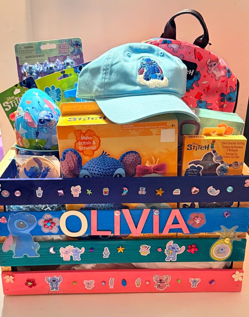 K&ouml;nnte beinhalten: Eine Holzkiste gef&uuml;llt mit Artikeln zum Thema Stitch. Enthalten sind eine hellblaue Baseballkappe mit einem gestickten Stitch-Design, ein rosa Rucksack, Bastelsets und Aufkleber. Der Name "OLIVIA" ist auf der Kiste zu sehen.