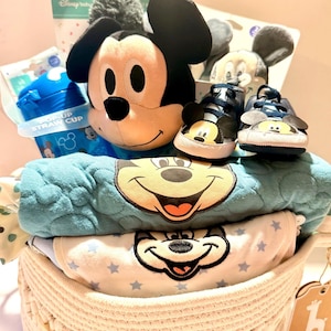 Op de afbeelding: Een baby cadeau mand met een Mickey Mouse knuffel, babyschoenen, een blauwe rietjesbeker en een blauwe deken met Mickey's gezicht. De mand bevat ook een wit en blauw rompertje met sterrenpatronen en een label met "Baby".