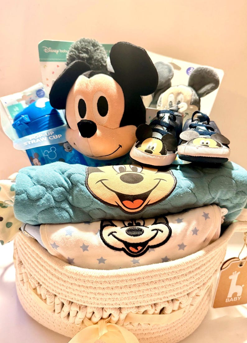 Op de afbeelding: Een baby cadeau mand met een Mickey Mouse knuffel, babyschoenen, een blauwe rietjesbeker en een blauwe deken met Mickey's gezicht. De mand bevat ook een wit en blauw rompertje met sterrenpatronen en een label met "Baby".