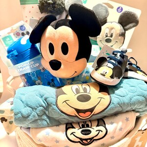 Op de afbeelding: Een Disney-thema babycadeaupakket gevuld met een Mickey Mouse knuffel, babyschoenen, een pop-up rietjesbeker en babykleding. De mand bevat een blauwe deken en een witte onesie met Mickey Mouse-ontwerpen. De mand is gemaakt van geweven materiaal.