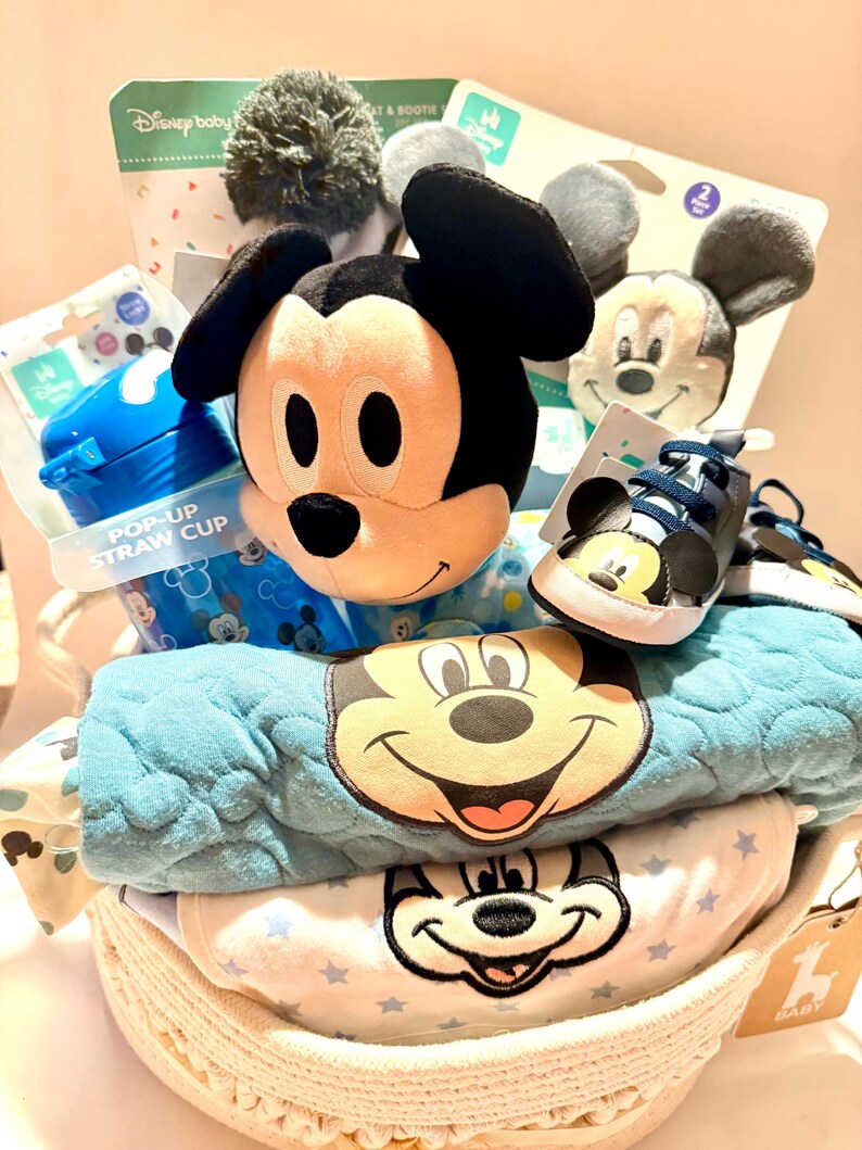 Op de afbeelding: Een Disney-thema babycadeaupakket gevuld met een Mickey Mouse knuffel, babyschoenen, een pop-up rietjesbeker en babykleding. De mand bevat een blauwe deken en een witte onesie met Mickey Mouse-ontwerpen. De mand is gemaakt van geweven materiaal.