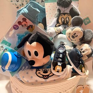 Op de afbeelding: Een babycadeaumand gevuld met Disney-artikelen. Inclusief een Mickey Mouse-knuffel, blauwe babyschoenen, een pop-up rietjesbeker, een muts, sokken en babykleding. De mand is wit en heeft een geweven ontwerp.
