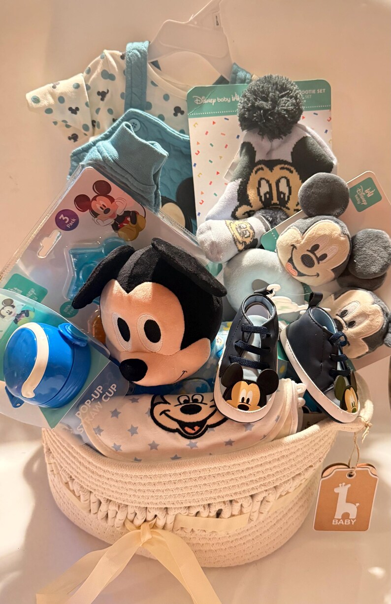 Op de afbeelding: Een babycadeaumand gevuld met Disney-artikelen. Inclusief een Mickey Mouse-knuffel, blauwe babyschoenen, een pop-up rietjesbeker, een muts, sokken en babykleding. De mand is wit en heeft een geweven ontwerp.