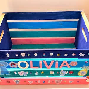 K&ouml;nnte beinhalten: Eine Holzkiste mit blauen, t&uuml;rkisfarbenen, rosa und goldenen Streifen. Die Kiste ist mit Aufklebern und dem Namen "OLIVIA" in gro&szlig;en rosa Buchstaben verziert. Sie hat Griffe an den Seiten.