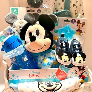 Op de afbeelding: Een Disney baby cadeau mand met Mickey Mouse thema items. Inclusief een Mickey knuffel, blauwe schoenen, een pop-up rietjesbeker, rammelaars, een muts en booties set en een kruipmaatje. De mand is wit en heeft een geweven ontwerp.