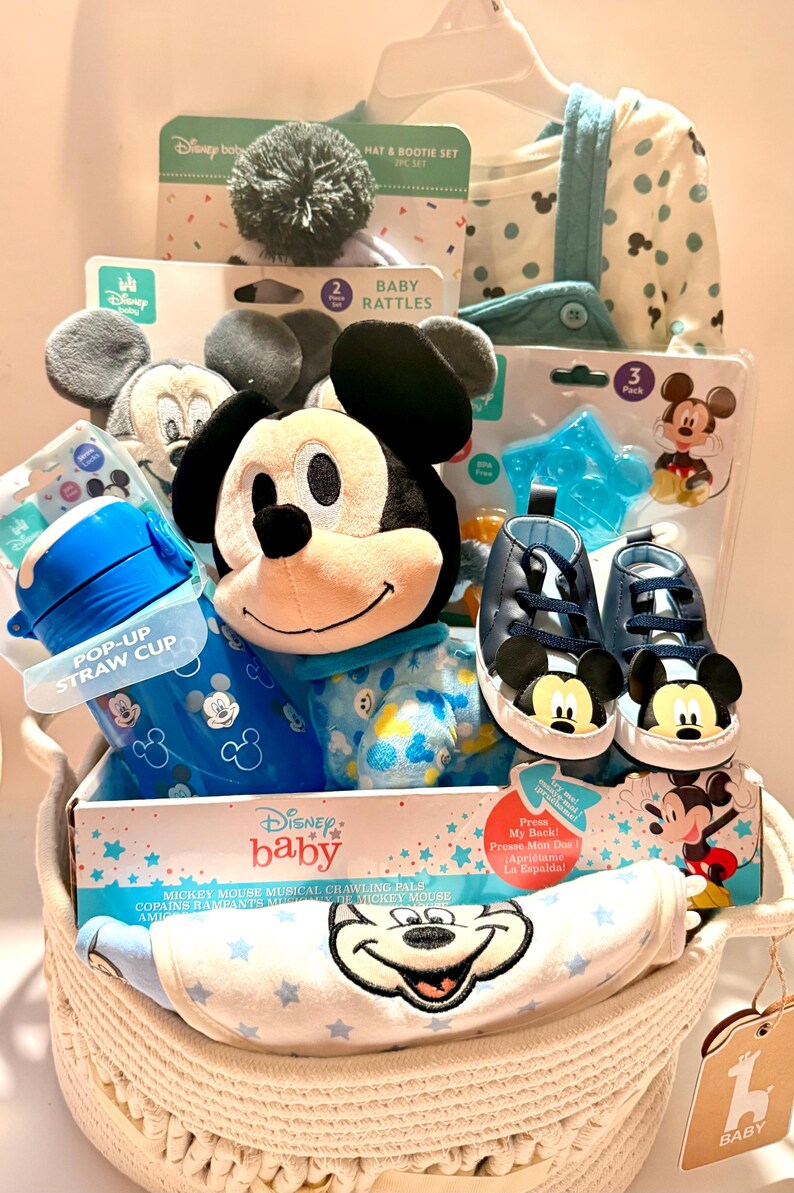 Op de afbeelding: Een Disney baby cadeau mand met Mickey Mouse thema items. Inclusief een Mickey knuffel, blauwe schoenen, een pop-up rietjesbeker, rammelaars, een muts en booties set en een kruipmaatje. De mand is wit en heeft een geweven ontwerp.