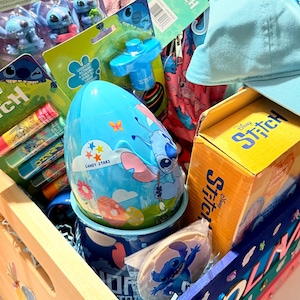 K&ouml;nnte beinhalten: Eine Holzkiste gef&uuml;llt mit Stitch-Artikeln. Sie enth&auml;lt eine blaue Baseballkappe, einen blauen eif&ouml;rmigen Beh&auml;lter mit dem Text "CANDY STARS", einen Becher, einen Keks und Spielzeug. Die Kiste ist mit bunten Akzenten und dem Namen "OLIVIA" verziert.