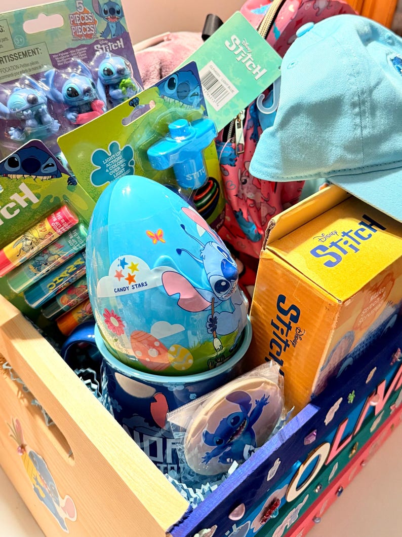 K&ouml;nnte beinhalten: Eine Holzkiste gef&uuml;llt mit Stitch-Artikeln. Sie enth&auml;lt eine blaue Baseballkappe, einen blauen eif&ouml;rmigen Beh&auml;lter mit dem Text "CANDY STARS", einen Becher, einen Keks und Spielzeug. Die Kiste ist mit bunten Akzenten und dem Namen "OLIVIA" verziert.