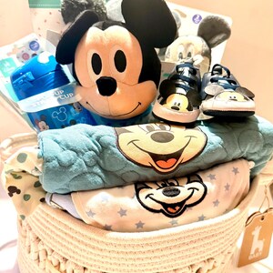 Op de afbeelding: Een gevlochten mand gevuld met babyartikelen. Inclusief een Mickey Mouse knuffel, blauwe babyschoenen, een blauwe tuitbeker en babykleding met Mickey Mouse-ontwerpen. De mand heeft een lichtbeige kleur en een gele lint.