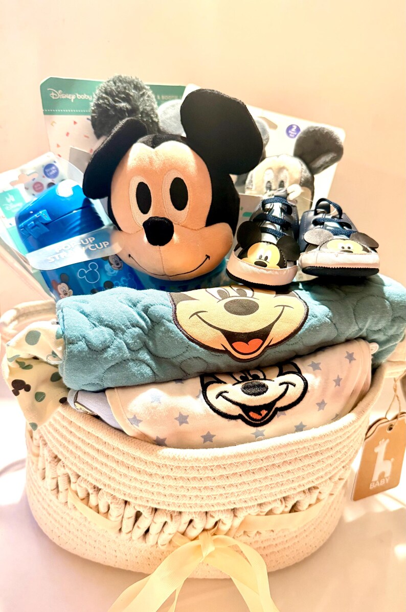 Op de afbeelding: Een gevlochten mand gevuld met babyartikelen. Inclusief een Mickey Mouse knuffel, blauwe babyschoenen, een blauwe tuitbeker en babykleding met Mickey Mouse-ontwerpen. De mand heeft een lichtbeige kleur en een gele lint.
