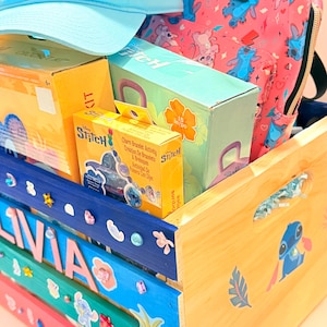 K&ouml;nnte beinhalten: Eine hellblaue Baseballkappe und ein pinkfarbener Rucksack mit der Figur Stitch sowie mehrere Kartons mit Stitch-Bastelsets befinden sich in einer Holzkiste. Die Kiste ist in verschiedenen Farben bemalt und mit Aufklebern und dem Namen "OLIVIA" verziert.